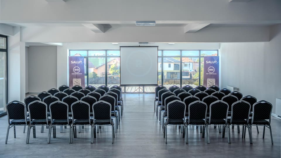 The hall laid out with chairs in rows, in two blocks, facing a projection screen, with tall windows running the width of the wall.